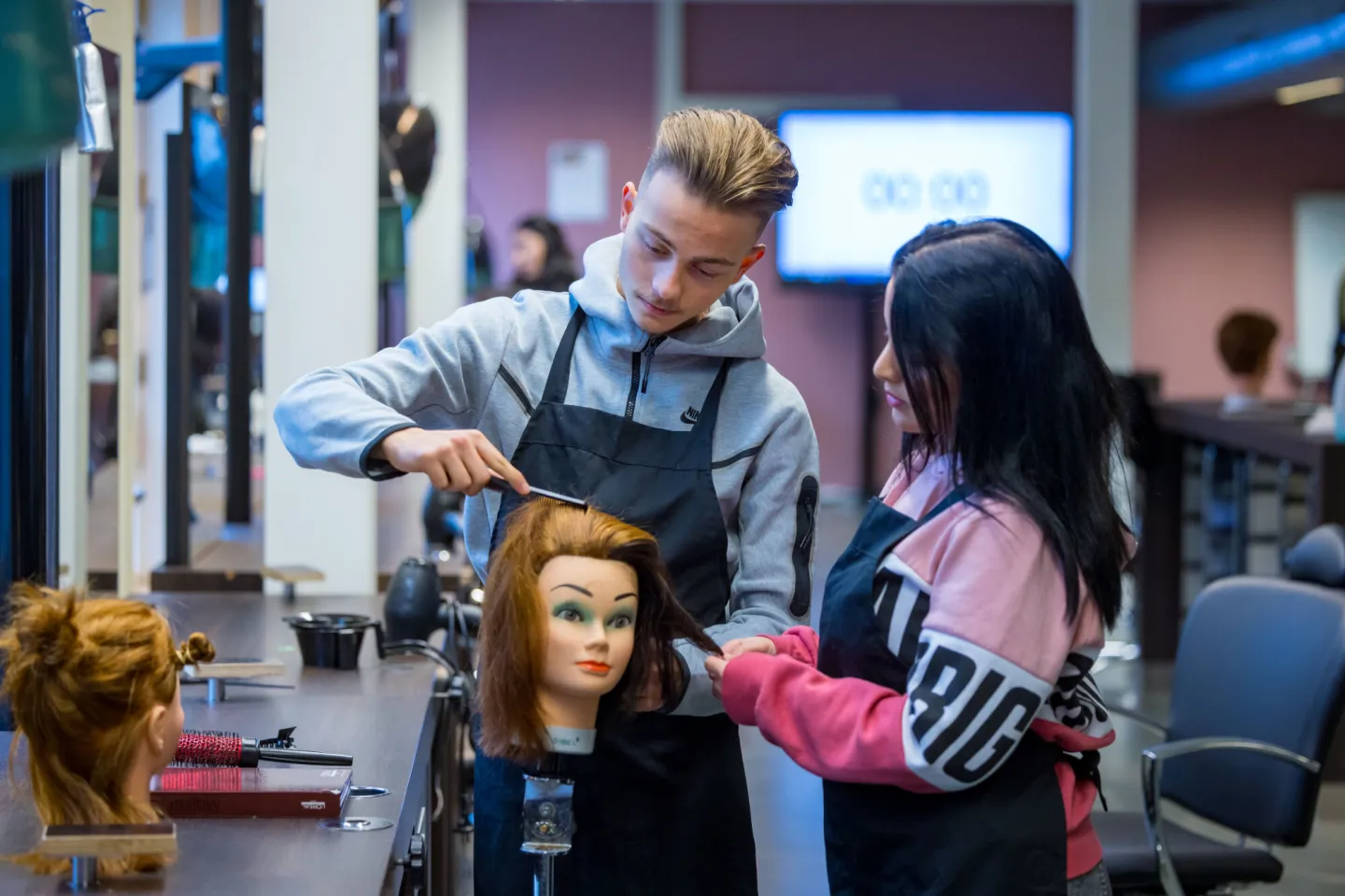 Twee personen knippen haar van een oefenpop in een salon.