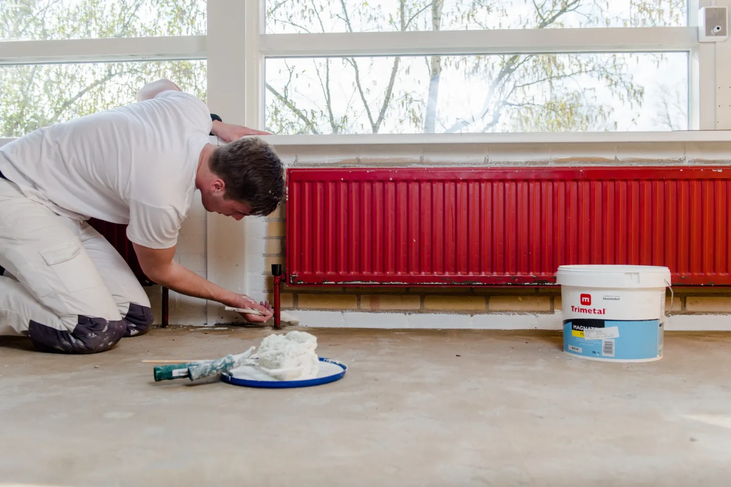 Een student schilder in werkkleding die een muur schildert met een rolborstel, omringd door verfblikken en gereedschap.