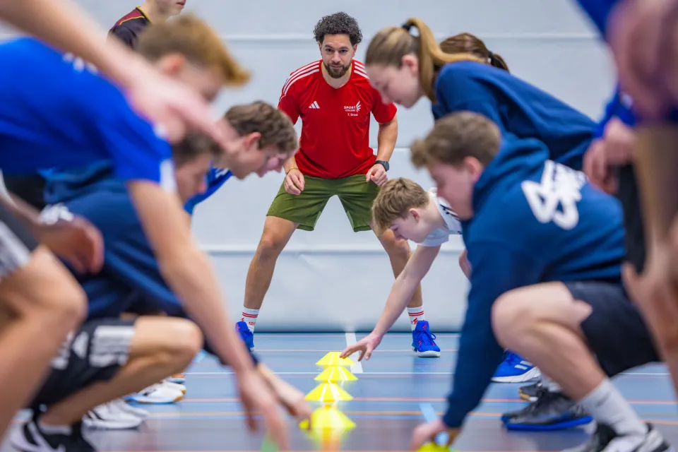 In een gymzaal zijn meerdere leerlingen met een oefening bezig. Achterin staat de docent aan te moedigen en te beoordelen hoe de leerlingen het uitvoeren. 