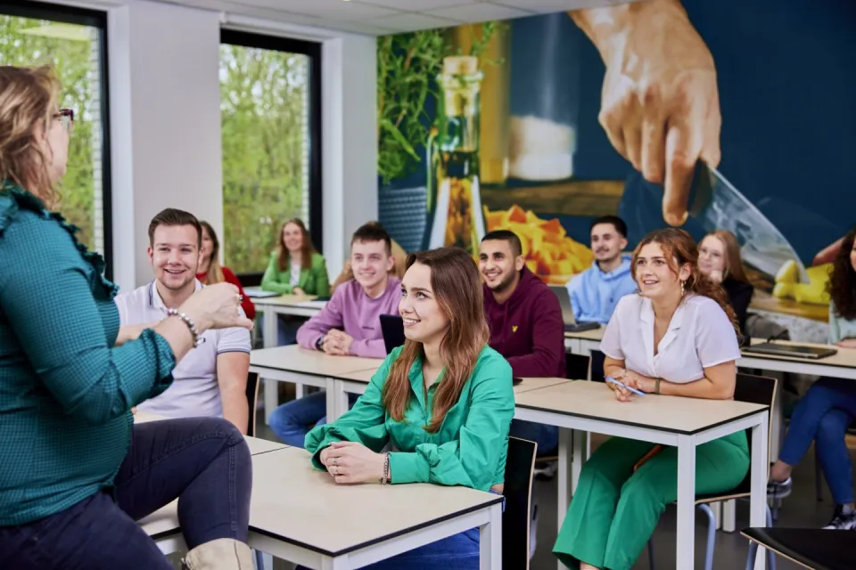 Een docent spreekt met een groep studenten in een klaslokaal, die aandachtig naar haar luisteren.