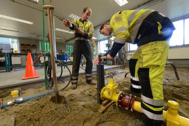 Twee monteurs gas-, water- en warmtedistributie werken aan een leiding op een bouwplaats. 