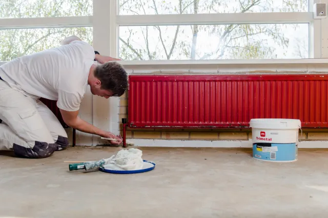 Een student Schilder schildert een radiator in een kamer met een verfkwast en verfblik.