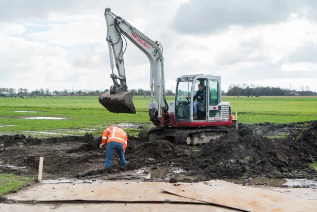 Waterbouwers aan het werk met graafmachine in weiland.