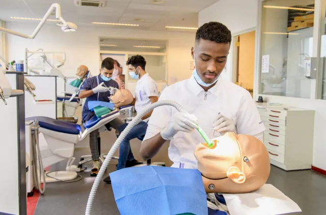 Een man werkt aan een pop in een tandartspraktijk. Op de achtergrond zie je ook twee studenten oefenen.