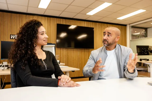 Een junior accountmanager en een klant zitten aan tafel en zijn in gesprek met elkaar.