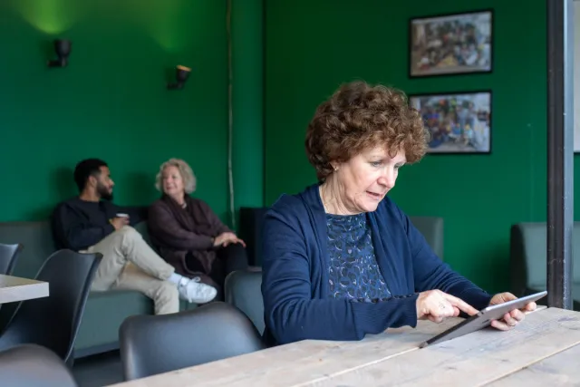 Vrouw bekijkt online zorgdossier op tablet in wachtkamer met op de achtergrond twee andere mensen. 