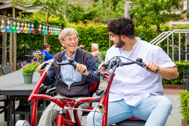 Een oudere man en een verzorger zitten samen op een speciale fiets voor twee personen. 