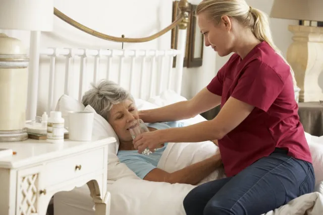 Verzorgende helpt oudere dame die in bed ligt bij drinken van glas water. 