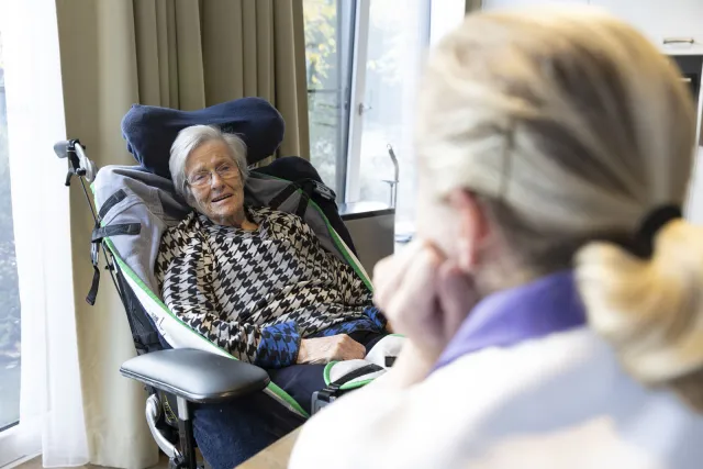 Een oudere vrouw in een rolstoel kijkt naar de zorgmedewerker tegenover haar.