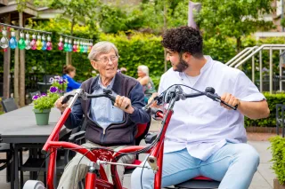 Een jonge man zit samen met een oudere patiënt op een tweezits-fiets. Ze glimlachen naar elkaar, hij is haar verzorger. 