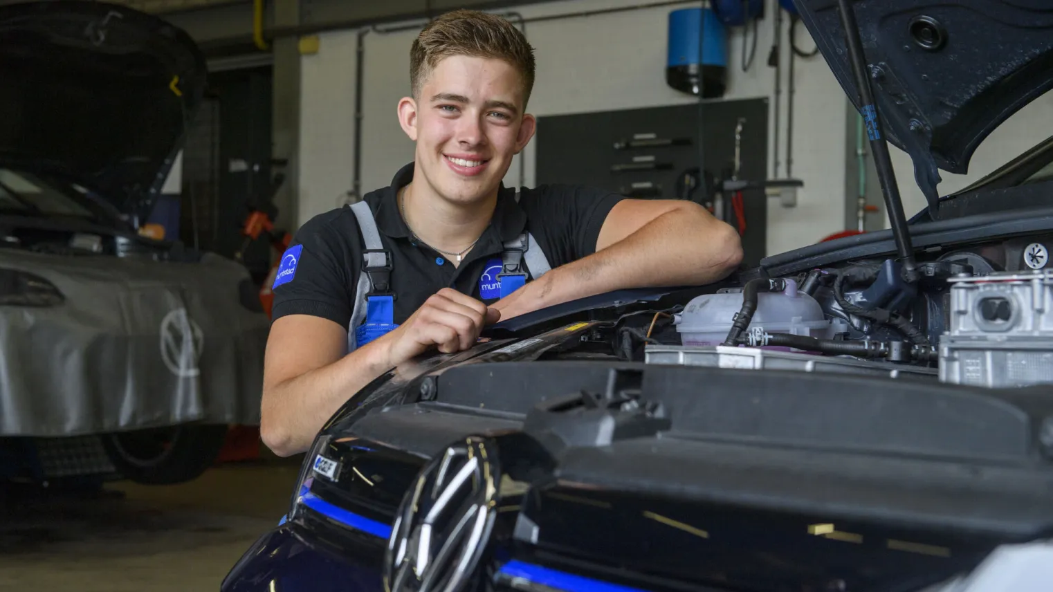 Student glimlacht terwijl hij voor een Volkswagen auto in de werkplaats zit.