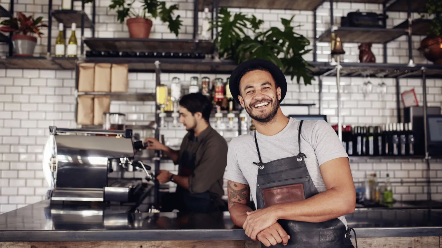 Barista in koffieshop leunt op de counter en achter hem maakt een collega koffie.