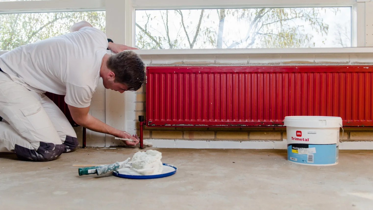 Een student Schilder schildert een radiator in een kamer met een verfkwast en verfblik.