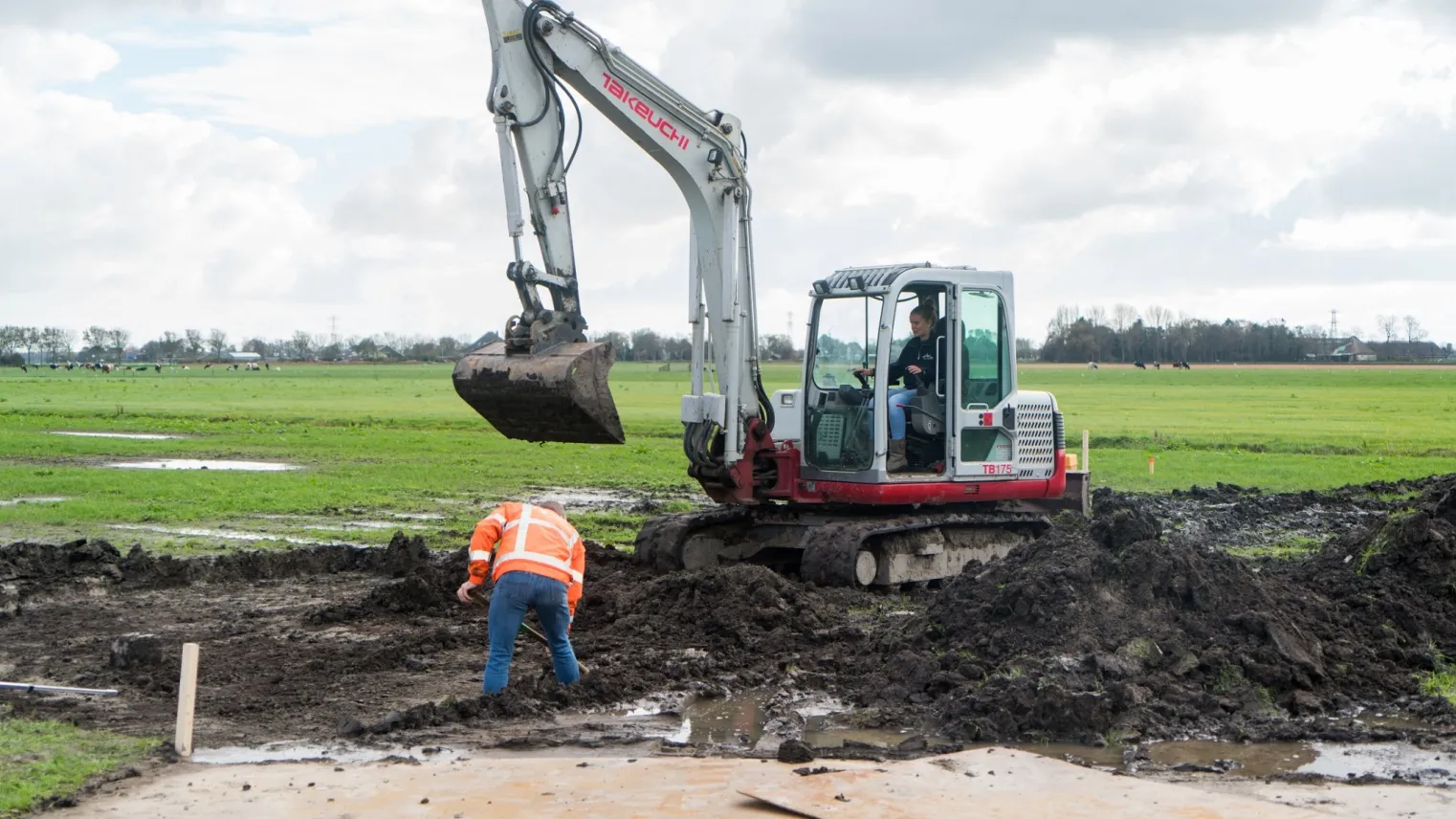 Waterbouwers aan het werk met graafmachine in weiland.