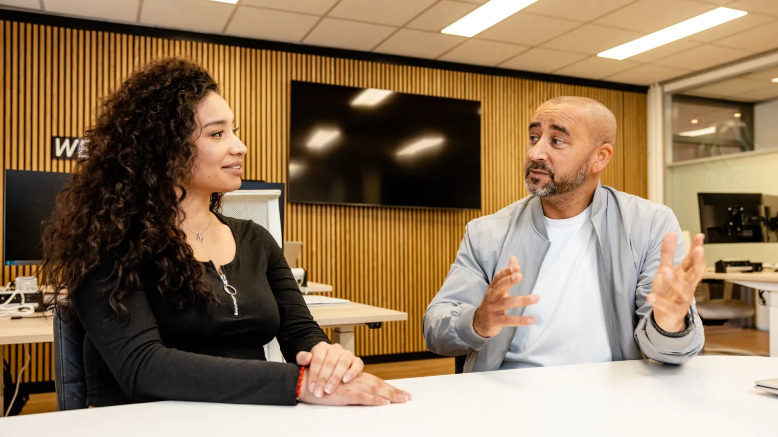 Een junior accountmanager en een klant zitten aan tafel en zijn in gesprek met elkaar.