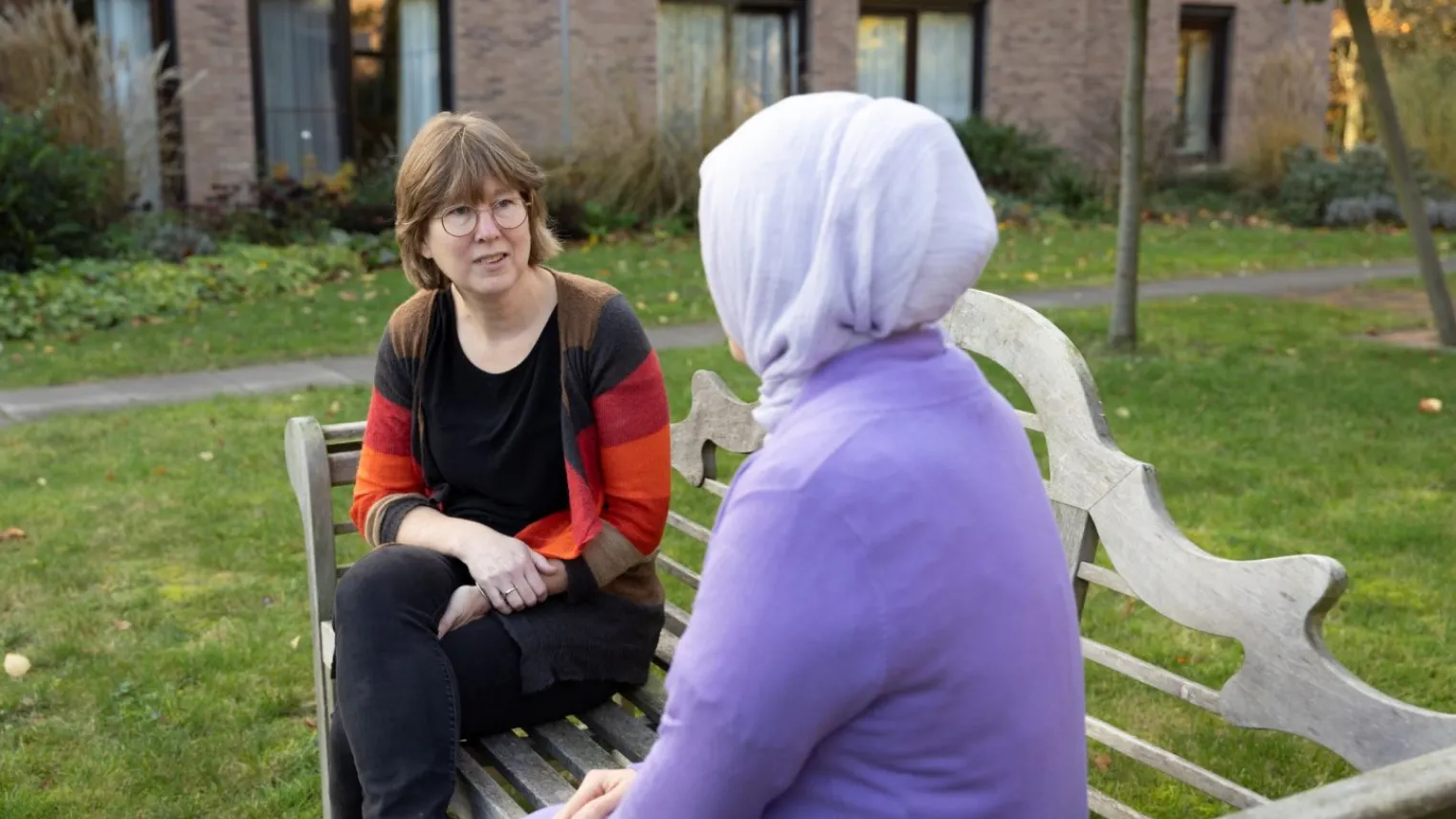 Praktijkopleider in gesprek met medewerker over leertraject buiten op bankje. 