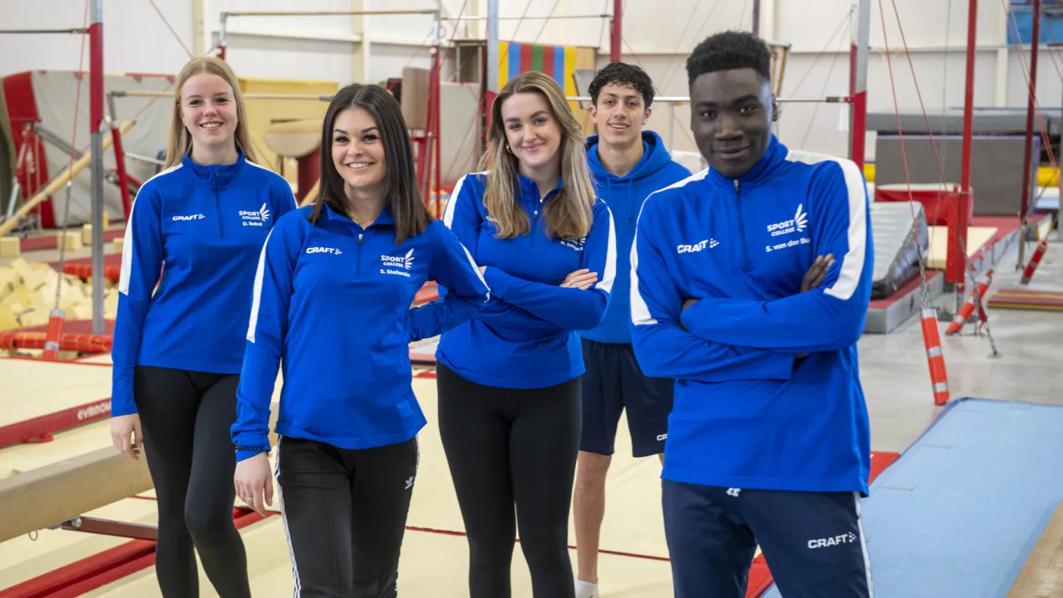 Vier jonge mensen in blauwe shirts en zwarte broeken staan op een gymnastiekvloer.