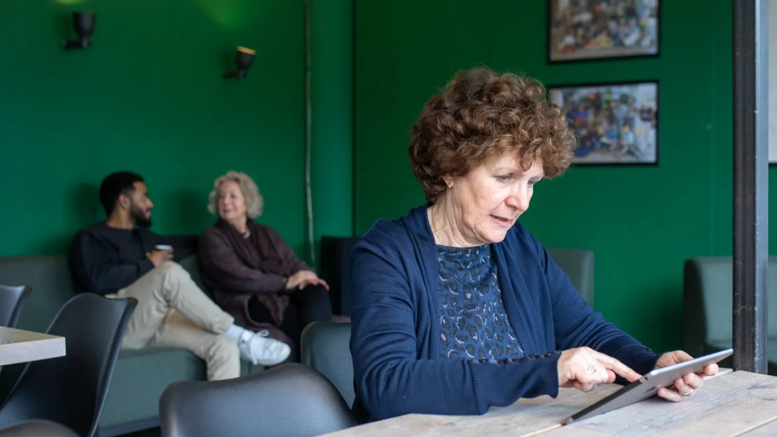 Vrouw bekijkt online zorgdossier op tablet in wachtkamer met op de achtergrond twee andere mensen. 