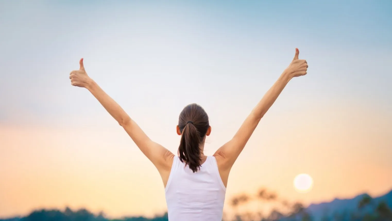 Vrouw staat met armen in de lucht en duimen omhoog bij zonsondergang. 