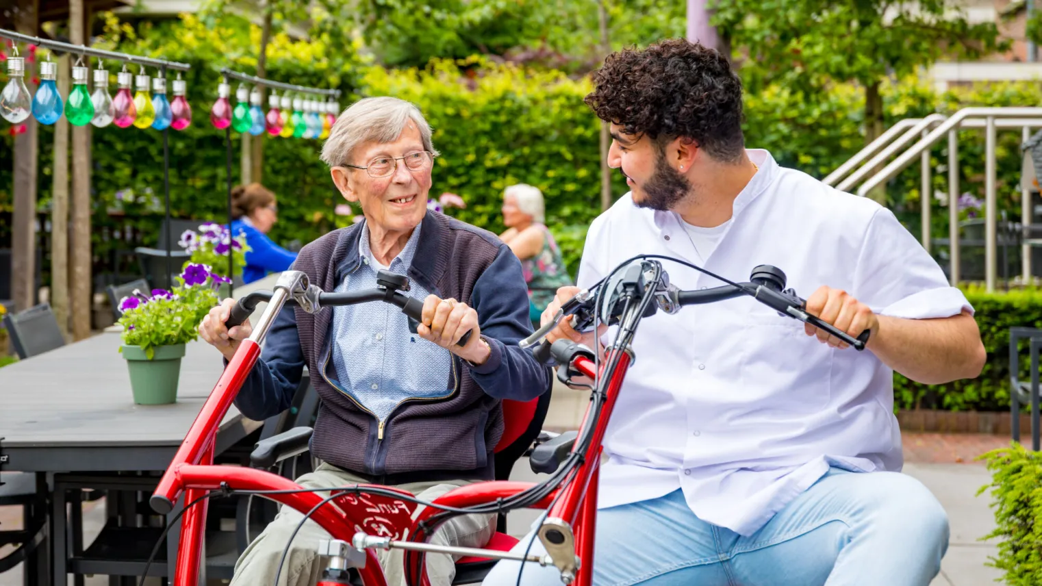 Een oudere man en een verzorger zitten samen op een speciale fiets voor twee personen. 