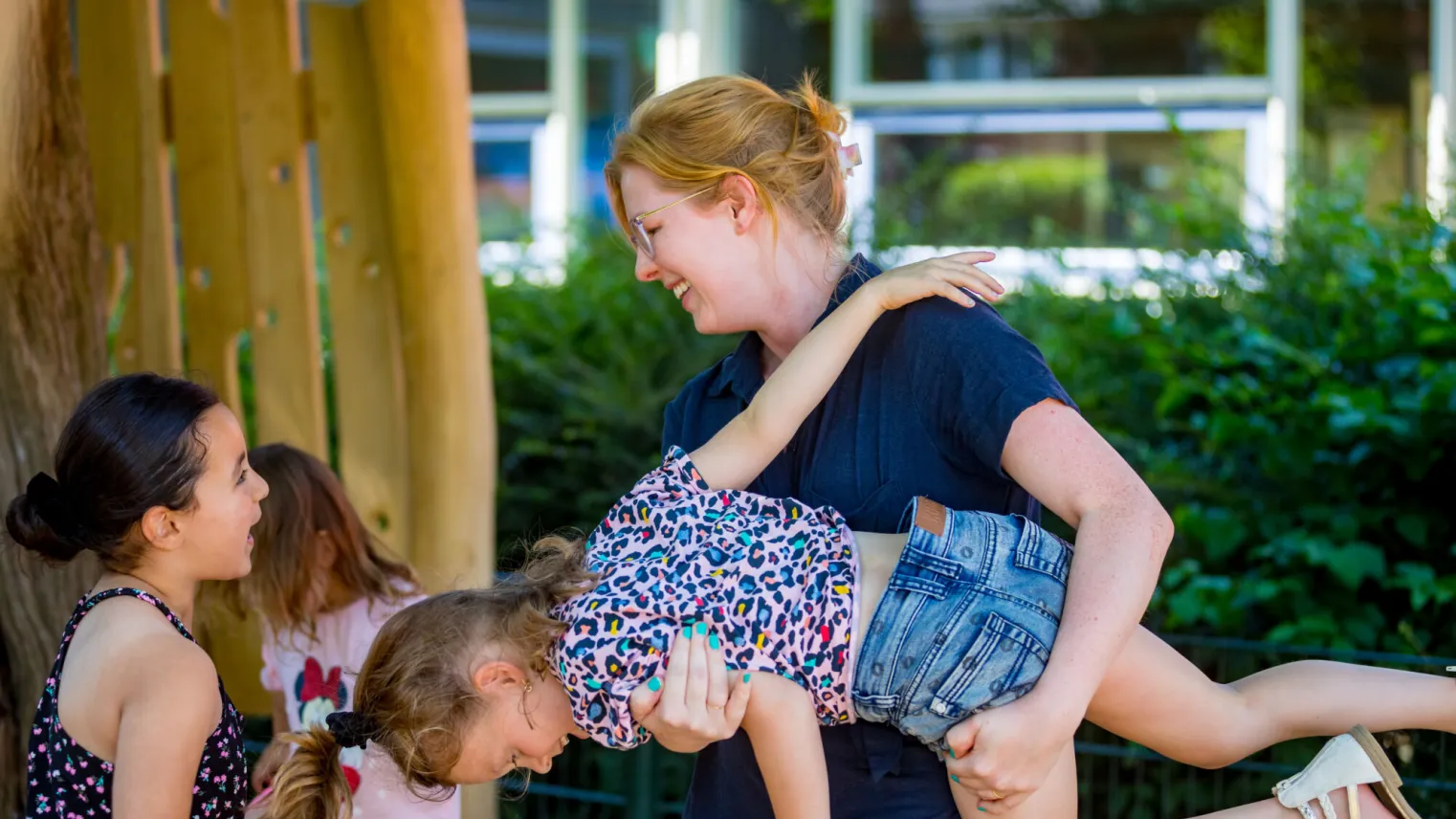 Een vrouw houdt een klein meisje in de lucht. Een vrolijke en speelse interactie tussen een begeleider en kinderen op een speelplaats. 