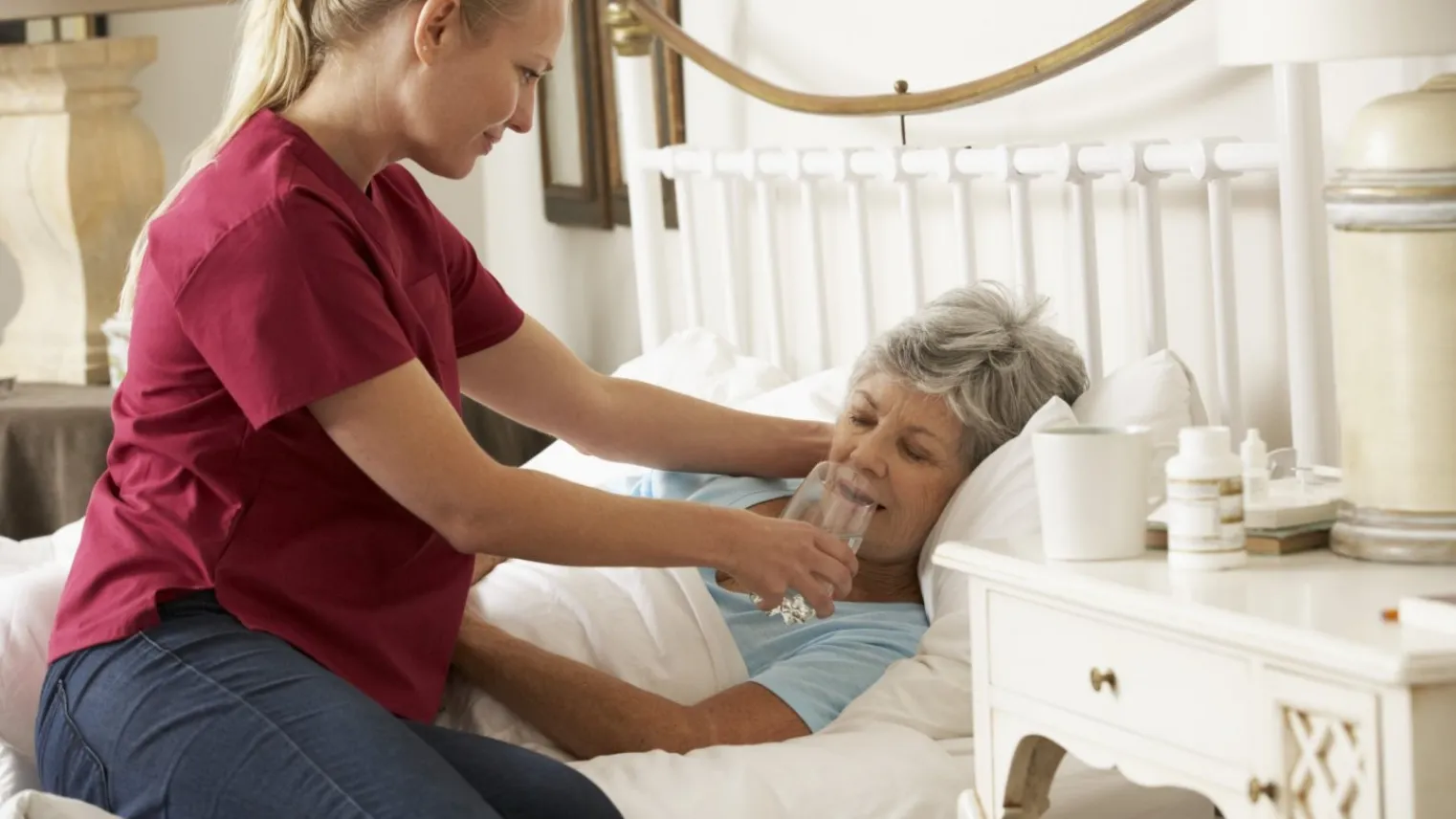 Verzorgende helpt cliënt die in bed ligt bij drinken van glas water. 