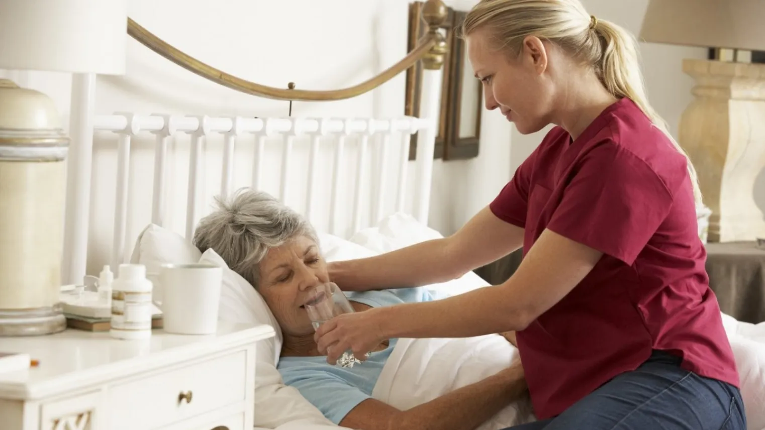 Verzorgende helpt oudere dame die in bed ligt bij drinken van glas water. 