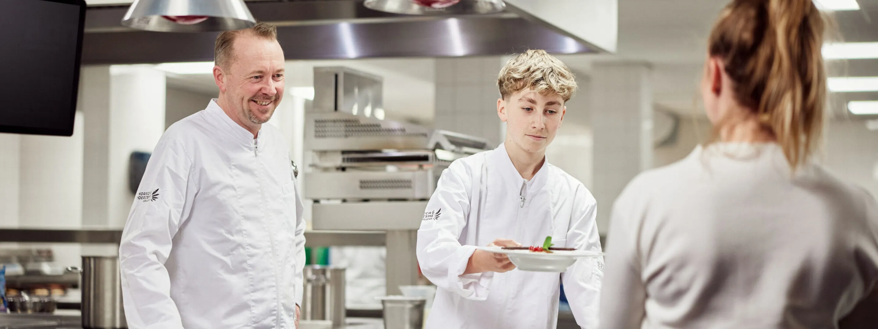 Een leerlingkok geeft vanuit de keuken een maaltijd aan een collega, onder het toeziend oog van de docent.