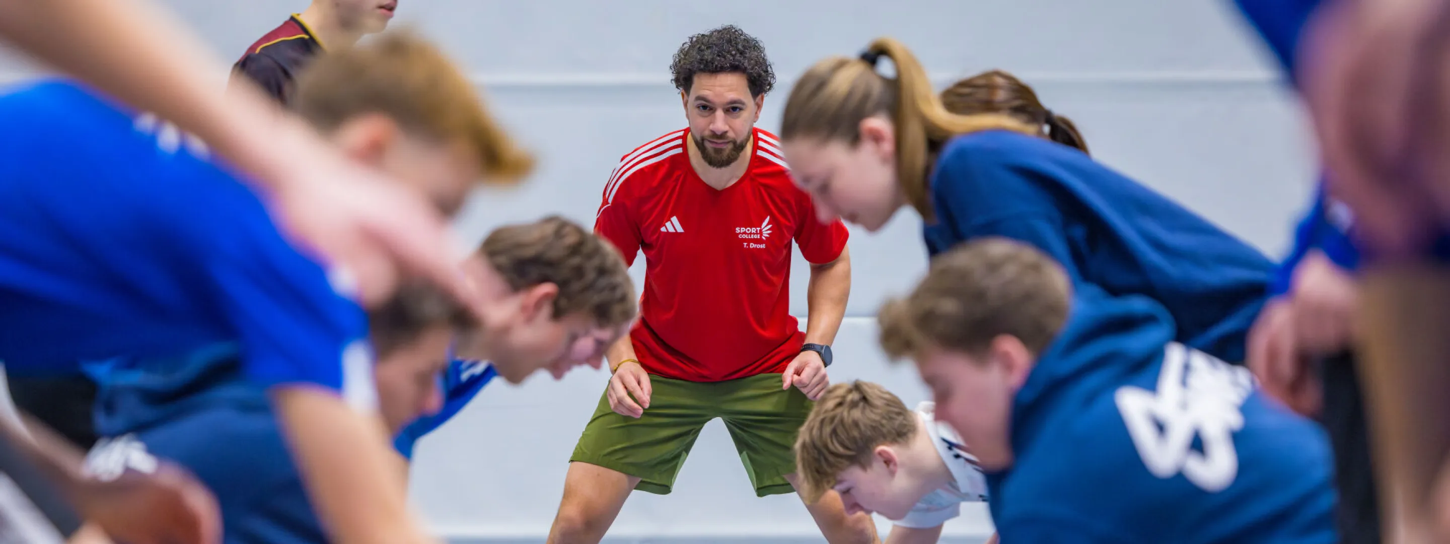 In een gymzaal zijn meerdere leerlingen met een oefening bezig. Achterin staat de docent aan te moedigen en te beoordelen hoe de leerlingen het uitvoeren. 