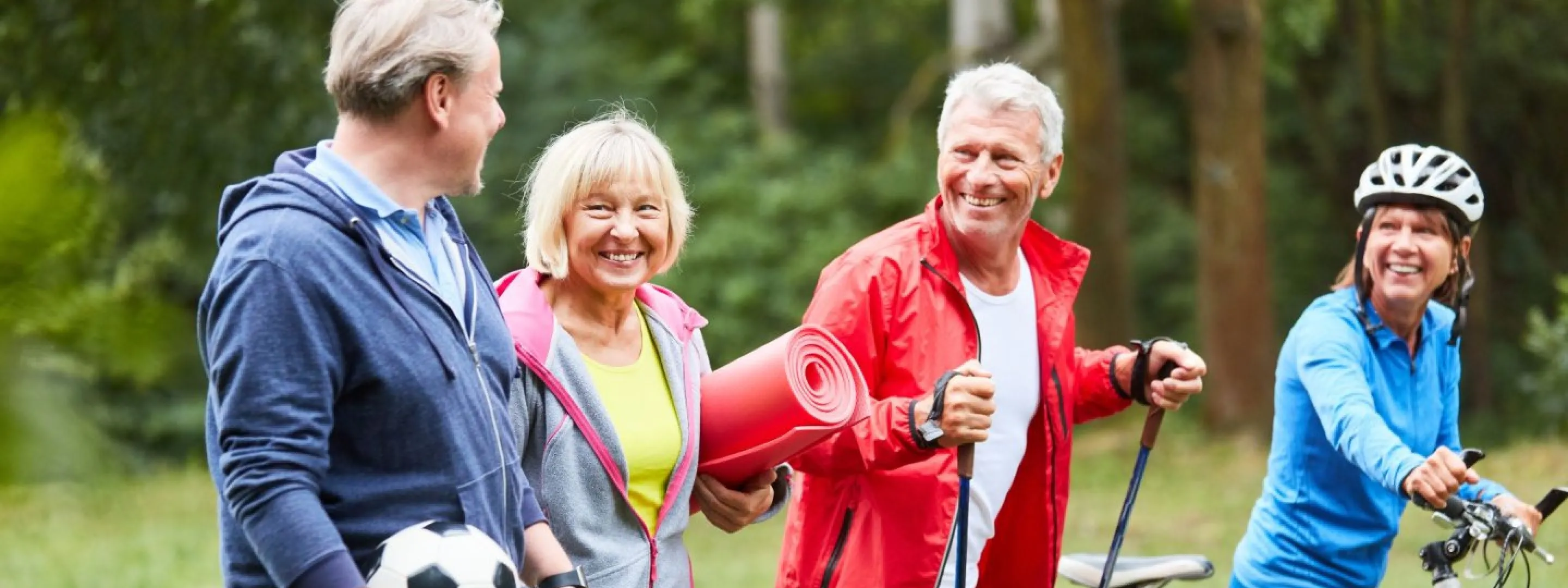 Vier volwassenen die sportief buiten zijn met voetbal, yogamat, Nordic walking sticks en een racefiets.