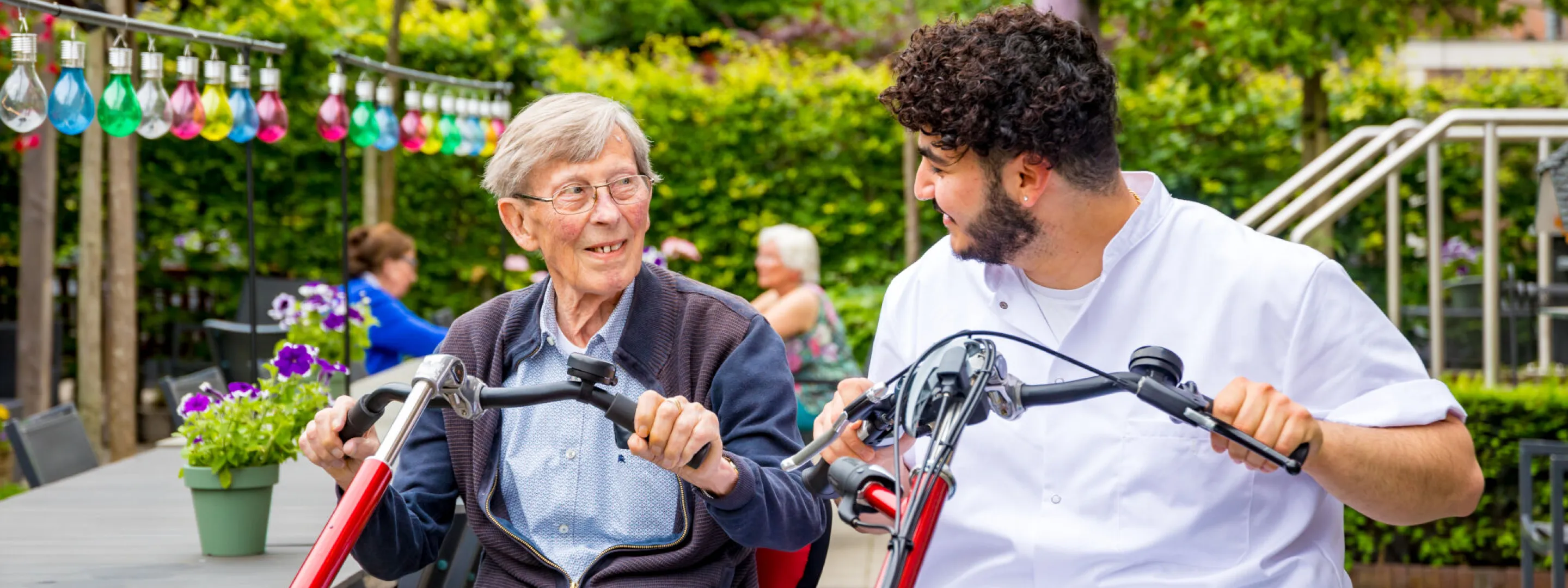 Een jonge man zit samen met een oudere patiënt op een tweezits-fiets. Ze glimlachen naar elkaar, hij is haar verzorger. 