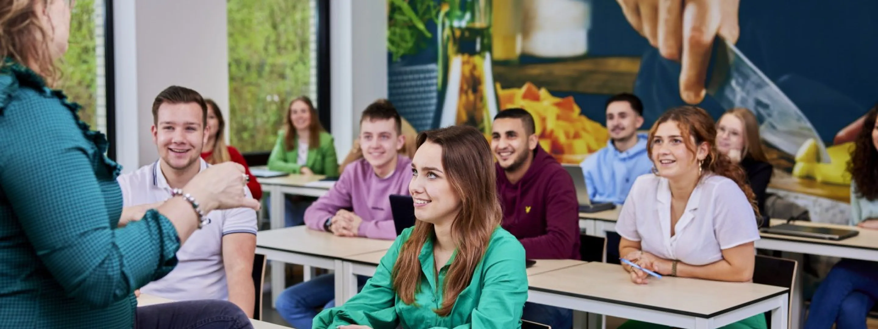 Een docent spreekt met een groep studenten in een klaslokaal, die aandachtig naar haar luisteren.